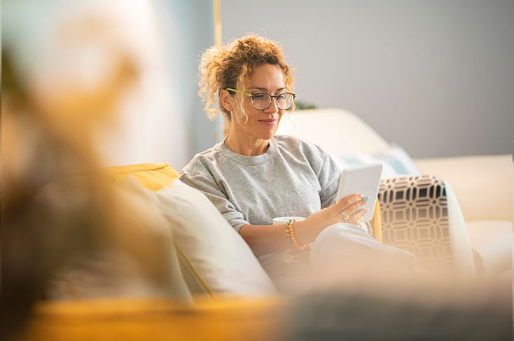 Frau lächelt - ein elektronisches Tablet in der Hand - auf dem Sofa.